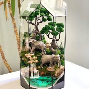 May include: A terrarium with a glass geometric frame, showcasing a miniature African savanna scene. It features two gray elephant figurines, green trees, moss, a waterfall, and a turquoise pond. The terrarium is a decorative piece.