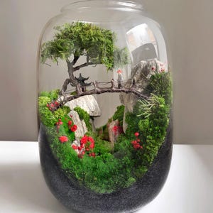 Könnte beinhalten: Eine Miniatur-Zen-Garten-Szene in einem transparenten Glas-Terrarium. Die Szene umfasst eine kleine Brücke, eine Pagode, einen Baum und rote Blumen. Der Sockel des Terrariums ist dunkelgrau.