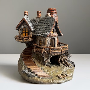 May include: A miniature fairy house sculpture with a stone base, wooden stairs, and a small balcony. The house features a gray shingled roof, wooden walls, and a chimney. The windows glow with a warm, inviting light.