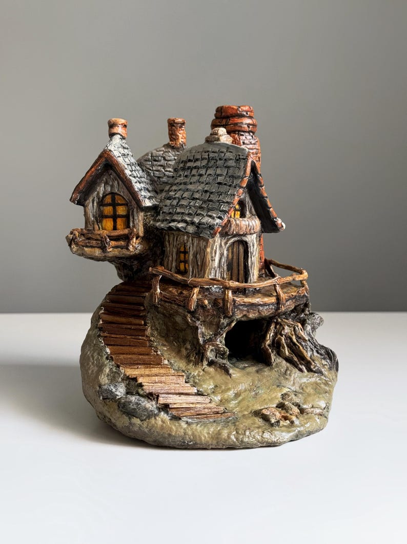 May include: A miniature fairy house sculpture with a stone base, wooden stairs, and a small balcony. The house features a gray shingled roof, wooden walls, and a chimney. The windows glow with a warm, inviting light.