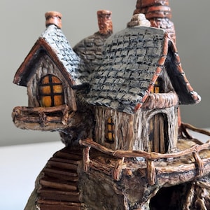 May include: A detailed miniature fairy house with a stone and wood exterior. The house features a gray shingled roof, small windows with yellow light, and a wooden door. A wooden staircase leads up to the house, and a small chimney is visible.