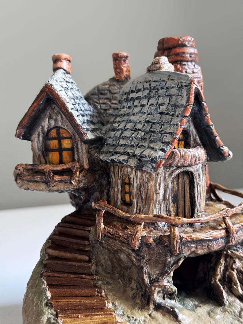 May include: A detailed miniature fairy house with a stone and wood exterior. The house features a gray shingled roof, small windows with yellow light, and a wooden door. A wooden staircase leads up to the house, and a small chimney is visible.