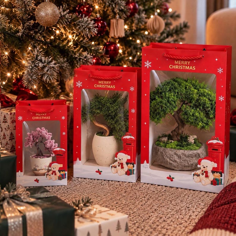 May include: Three red gift bags with clear windows, each containing a small potted tree. The bags feature the text Merry Christmas, a red mailbox, and a teddy bear. The smallest bag holds a pink tree, while the larger bags hold green trees.