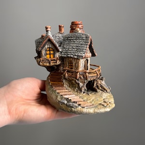 May include: A miniature fantasy house figurine with a stone base, wooden stairs, and a balcony. The house has a gray shingled roof, a chimney, and windows with a warm, inviting glow. The figurine is held in a hand.