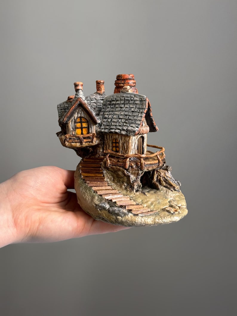 May include: A miniature fantasy house figurine with a stone base, wooden stairs, and a balcony. The house has a gray shingled roof, a chimney, and windows with a warm, inviting glow. The figurine is held in a hand.