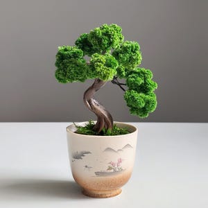 May include: A small bonsai tree in a decorative ceramic pot. The tree has bright green, textured foliage and a curved, brown trunk. The pot is beige with a landscape design featuring mountains and a small tree with pink flowers.