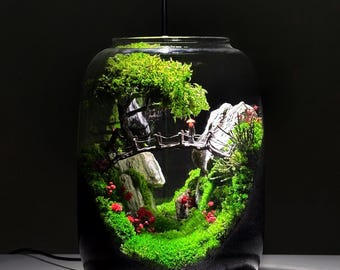 Op de afbeelding: Een miniatuur terrarium in een glazen pot, met een landschap met een brug, bomen en rode bloemen. De scène wordt verlicht door een kleine spot, wat een dramatisch effect creëert. De pot staat op een donkere basis.