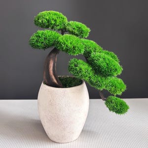Pode incluir: Um bonsai artificial com um tronco curvo castanho-escuro e aglomerados de folhagem verde vibrante em forma de agulha. A árvore está plantada num vaso de cerâmica oval, de cor esbranquiçada e texturizada, com uma base coberta de musgo.