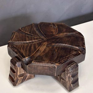 Puede incluir: Un taburete bajo de madera con un acabado teñido en marrón oscuro. La parte superior circular presenta ranuras talladas que irradian desde el centro, y la pieza se apoya en tres patas gruesas y robustas con detalles tallados en los laterales.