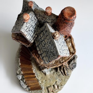 May include: A miniature, hand-painted ceramic village scene. The piece features multiple gray-roofed buildings, a brown chimney, and a wooden staircase. The base is textured with a stone-like appearance.