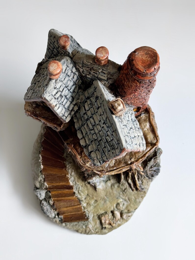 May include: A miniature, hand-painted ceramic village scene. The piece features multiple gray-roofed buildings, a brown chimney, and a wooden staircase. The base is textured with a stone-like appearance.