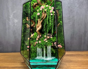 May include: A terrarium with a glass geometric prism shape. Inside, a miniature landscape features a waterfall, moss, and a wooden sculpture of a figure. Pink flowers and green foliage add color to the scene. The base has a turquoise-colored water feature.