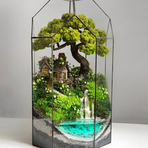 Puede incluir: Un terrario en miniatura con una estructura de vidrio. En su interior, un paisaje detallado presenta una pequeña cabaña, una cascada y un estanque con agua turquesa. Plantas verdes exuberantes y un gran árbol completan la escena.