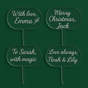 May include: Four clear acrylic cake toppers with rounded oval tops and pointed stems. Each topper has white script text: "With love, Emma", "Merry Christmas, Jack", "To Sarah, with magic", and "Love always, Noah & Lily". The background is a dark green.
