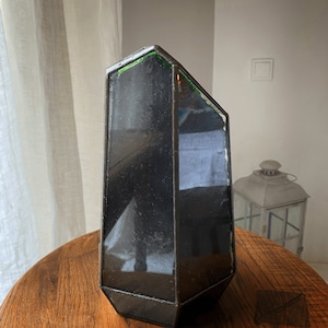 Op de afbeelding: Een zwarte geometrische glazen kaarsenhouder met een groene glazen accent. De houder staat op een houten tafel.