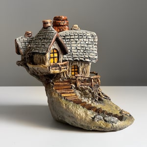 May include: A miniature, whimsical cottage sculpture. The house features a gray shingled roof, a stone chimney, and a warm, inviting glow from the windows. A wooden staircase leads up to the cottage, which is set on a rocky base.