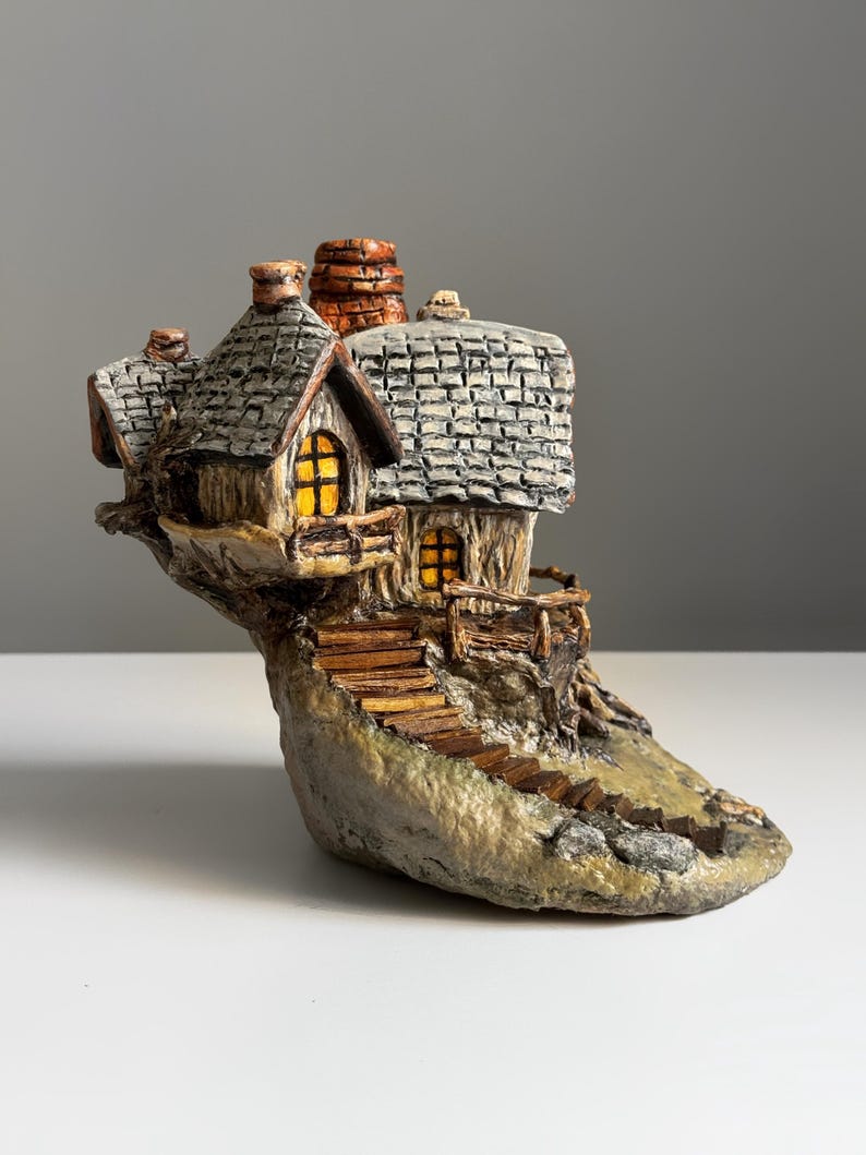 May include: A miniature, whimsical cottage sculpture. The house features a gray shingled roof, a stone chimney, and a warm, inviting glow from the windows. A wooden staircase leads up to the cottage, which is set on a rocky base.