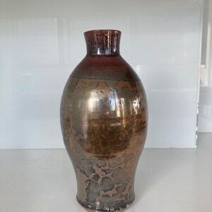 Peut inclure: Un vase en céramique avec un glaçage brun, or et noir. Le vase a un corps arrondi et un col étroit.