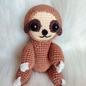 May include: A brown crocheted sloth with white crocheted feet and black eyes. The sloth is sitting on a white fluffy surface.