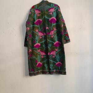 May include: A long, dark green coat with a floral pattern. The coat features large pink flowers with teal stems and leaves. The coat is hanging on a black hanger against a white wall.