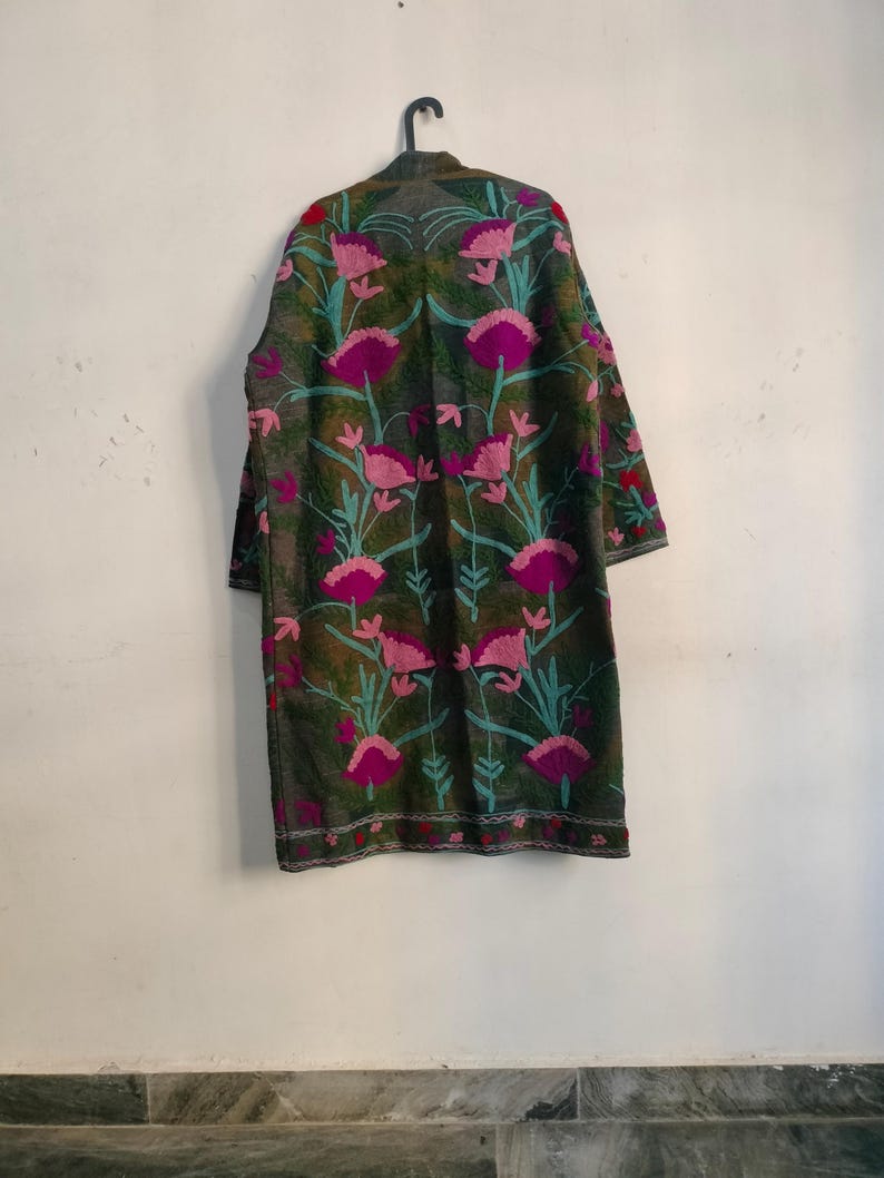 May include: A long, dark green coat with a floral pattern. The coat features large pink flowers with teal stems and leaves. The coat is hanging on a black hanger against a white wall.
