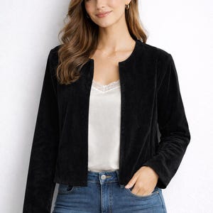 Può includere: Giacca nera in velluto senza colletto. La giacca è aperta sul davanti, rivelando una camisole bianca con finiture in pizzo. La modella indossa jeans blu. L'aspetto generale è casual e chic.