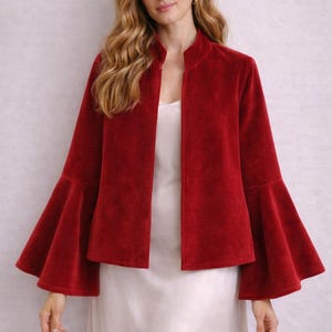 Puede incluir: Chaqueta de terciopelo rojo con cuello alzado y mangas acampanadas. La chaqueta es abierta por delante y se lleva sobre un vestido blanco. Las mangas son anchas y acampanadas, lo que añade un toque dramático a la prenda.