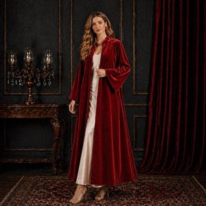 Puede incluir: Una larga bata de terciopelo rojo intenso con cuello alto, sobre un vestido color crema. El fondo presenta paredes oscuras, una alfombra estampada y un candelabro decorativo.