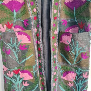 May include: A long, open-front jacket with intricate floral embroidery. The jacket features large, stylized flowers in shades of pink and purple, with teal green stems and leaves. The embroidery is set against a muted olive green and gray background.