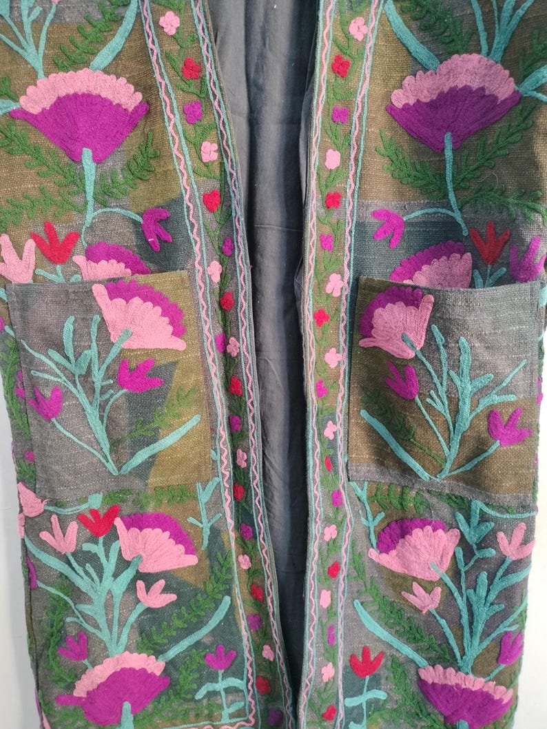 May include: A long, open-front jacket with intricate floral embroidery. The jacket features large, stylized flowers in shades of pink and purple, with teal green stems and leaves. The embroidery is set against a muted olive green and gray background.