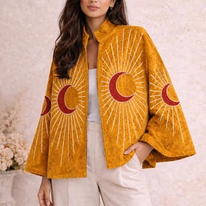 Puede incluir: Chaqueta color mostaza con diseño celestial. La chaqueta tiene cuello alto y mangas anchas. La parte delantera y las mangas están adornadas con una media luna roja y bordados de rayos de sol blancos. La modelo lleva un top blanco y pantalones color crema.