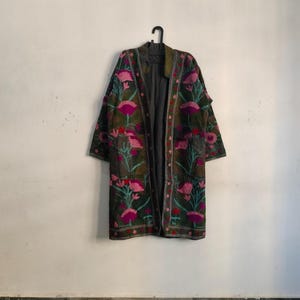May include: A long, open-front coat with a dark olive green base, adorned with vibrant pink and teal floral embroidery. The coat features a dark gray interior and is hanging on a black hanger against a white wall.