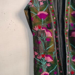 May include: An open-front, embroidered jacket with a floral design. The jacket features a dark green base with patches of brown and gray. The embroidery includes pink, purple, and teal flowers with red accents. The jacket has a relaxed fit.