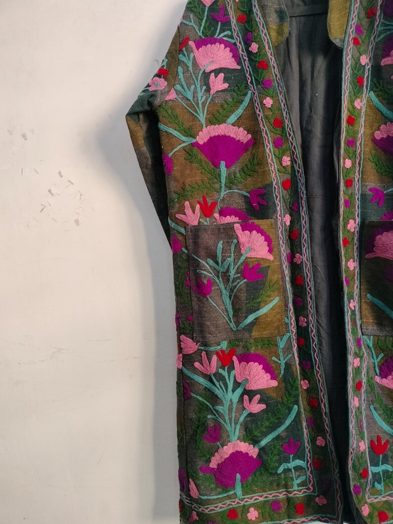 May include: An open-front, embroidered jacket with a floral design. The jacket features a dark green base with patches of brown and gray. The embroidery includes pink, purple, and teal flowers with red accents. The jacket has a relaxed fit.