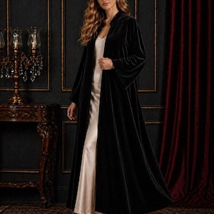 Pode incluir: Um robe longo de veludo preto com gola xale e mangas compridas, usado sobre um vestido de cetim creme. O fundo inclui uma parede escura, uma cortina vermelha e um tapete estampado.
