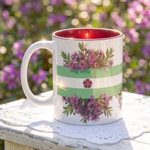 May include: White ceramic mug with a red interior and a white handle. The mug features a design of purple flowers and green stripes. A single purple flower is centered on the mug. The background is a blurred view of purple flowers.