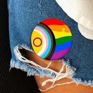 May include: A round button with a rainbow design and a heart. The button also features the colors of the transgender flag and the intersex flag. The button is held against a denim background.