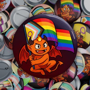 Puede incluir: Una colección de botones coloridos con un diablo de dibujos animados sosteniendo una bandera del orgullo arcoíris. El diablo es naranja con cuernos rojos y una llama amarilla en la cabeza. Los botones están rodeados de otras banderas y símbolos del orgullo.