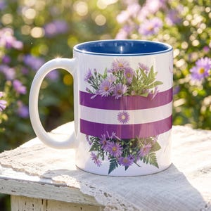May include: White ceramic mug with a blue interior and a handle. The mug features a design of purple flowers and horizontal purple stripes. The background is a blurred view of purple flowers.