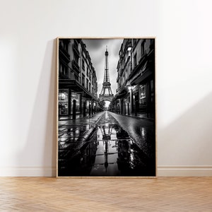 Puede incluir: Una fotografía en blanco y negro de la Torre Eiffel en París, Francia. La torre se ve desde una perspectiva a nivel de calle, con edificios que bordean la calle a ambos lados. La calle está mojada por la lluvia, y el reflejo de la torre se puede ver en un charco.