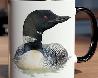 May include: A white ceramic mug with a black handle and rim. The mug features a watercolor illustration of a loon floating on water. The loon has black and white feathers, a black head, and a red eye. The mug is on a wooden surface.