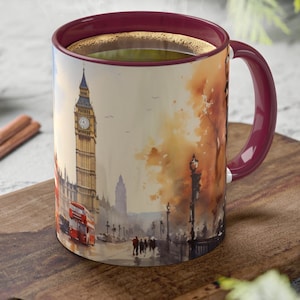 May include: A white ceramic mug with a watercolor image of Big Ben and a red double-decker bus in London, England. The mug has a burgundy rim and handle. The mug is sitting on a wooden cutting board.