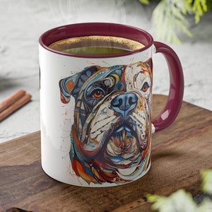 May include: A white ceramic mug with a burgundy interior and handle, featuring a colorful, artistic illustration of a bulldog's face. The mug is filled with a dark liquid, possibly coffee or tea, and is placed on a wooden surface.