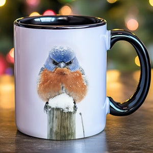May include: A white ceramic coffee mug with a black rim and handle. The mug features a colorful image of a bluebird perched on a wooden post, covered in snow.