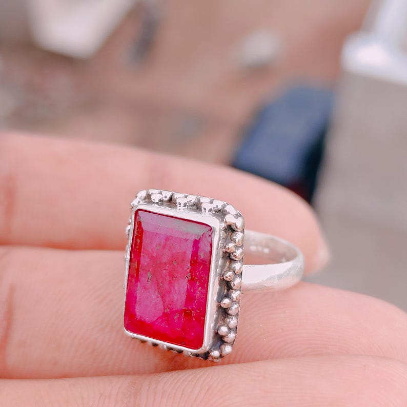 May include: A silver ring featuring a large, rectangular, faceted hot pink gemstone set in a bezel. The metal setting is decorated with a border of small silver spheres, and the band has a smooth, polished finish with a simple, rounded design.