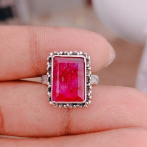 May include: A silver ring featuring a rectangular, faceted, vibrant pink stone set in a decorative, beaded metal frame. The stone displays natural internal veining and texture, while the band is crafted from a polished, metallic material.