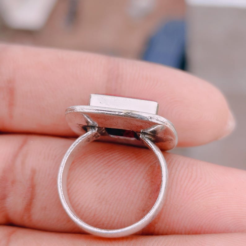 May include: A silver-toned ring featuring a rectangular setting with a flat, reddish-orange stone inlay. The metal band is circular and has a slightly weathered, textured finish around the base of the rectangular stone mount.