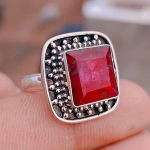 May include: A silver ring featuring a square-cut, deep red gemstone set in a frame decorated with small metal beads. The setting has a dark, oxidized finish around the stone, contrasting with the polished silver band and the textured bead border.