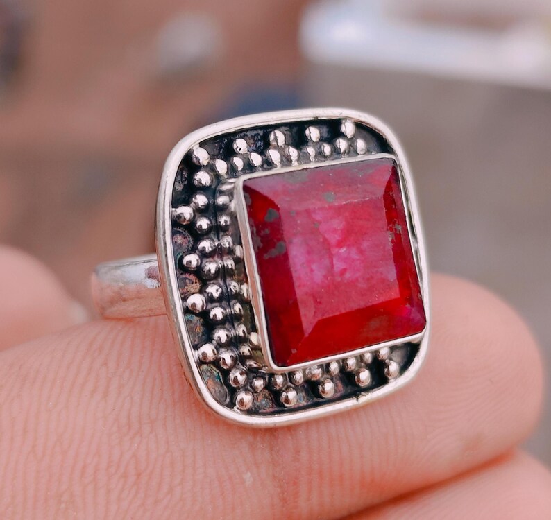 May include: A silver ring featuring a square-cut, deep red gemstone set in a frame decorated with small metal beads. The setting has a dark, oxidised finish around the stone, contrasting with the polished silver band and the textured bead border.