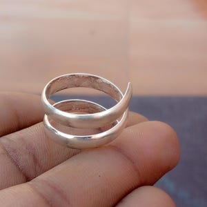 Könnte beinhalten: Ein Ring aus silberfarbenem Metall mit einem Bypass-Design, bei dem zwei überlappende Bänder spiralförmig um den Finger verlaufen. Die Oberfläche des Rings hat ein glattes, mattes Finish mit abgerundetem Profil und offener Kreisform.